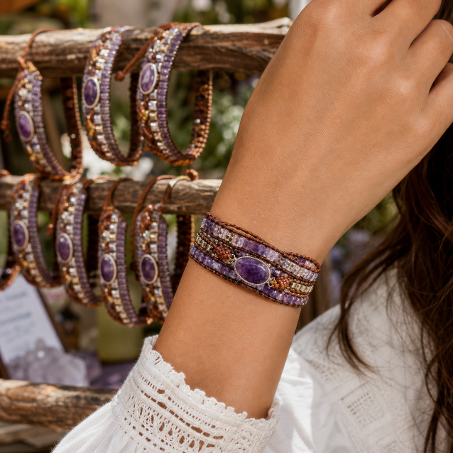 A close-up of a person's wrist wearing a multi-strand beaded bracelet with purple amethyst stones and brown and gold beads, with other matching bracelets hanging on a rustic wooden stand in the background.